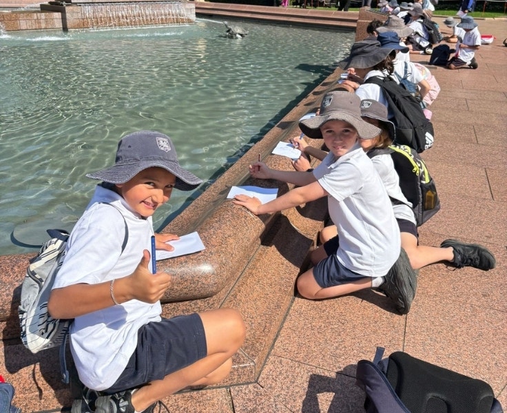 Students are sitting at a fountain. Two are facing the camera. One is giving a thumbs up. The students have paper in front of them.