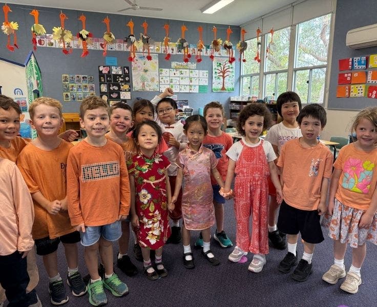 A Kindergarten class, wearing orange clothes, are standing in a line and smiling at the camera.