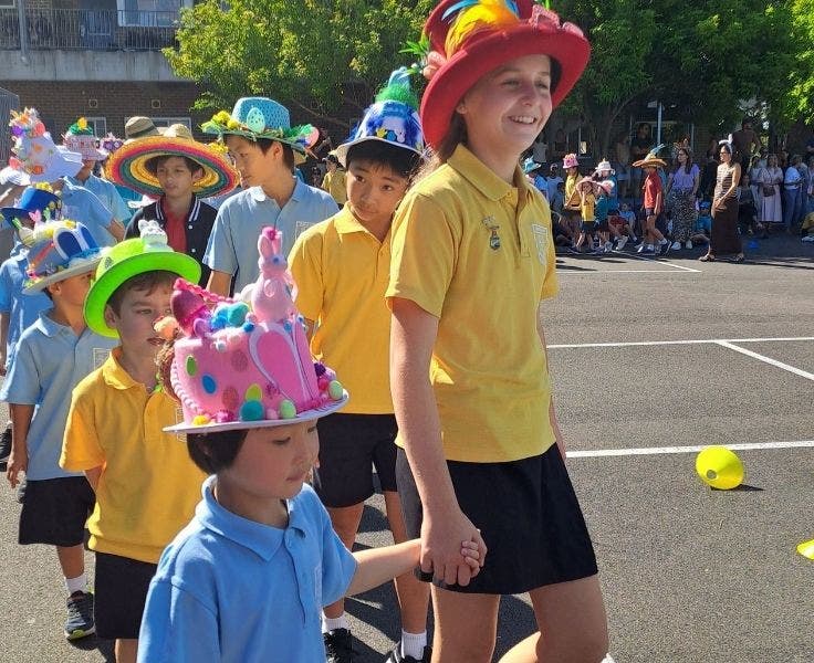 A Senior student wearing a yellow shirt and red hat walking with her Kindergarten buddy wearing a blue shirt and a pink hat. Two lines of students are following them.