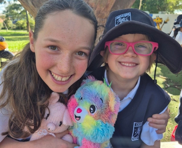 A Year 6 student sitting next to and hugging her Kindergarten buddy. They are both holding cuddly toys and smiling.