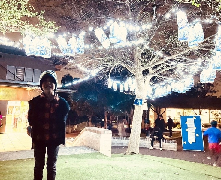A male student standing in front of a tree that has string lights illuminating the tree and the plastic bottles attached to highlight the plastic waste problem.