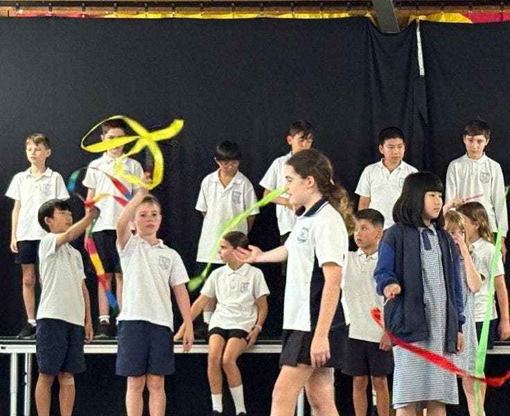 A female student is walking across the stage in front of a group of students. Some of the students in the background are waving colourful ribbons.