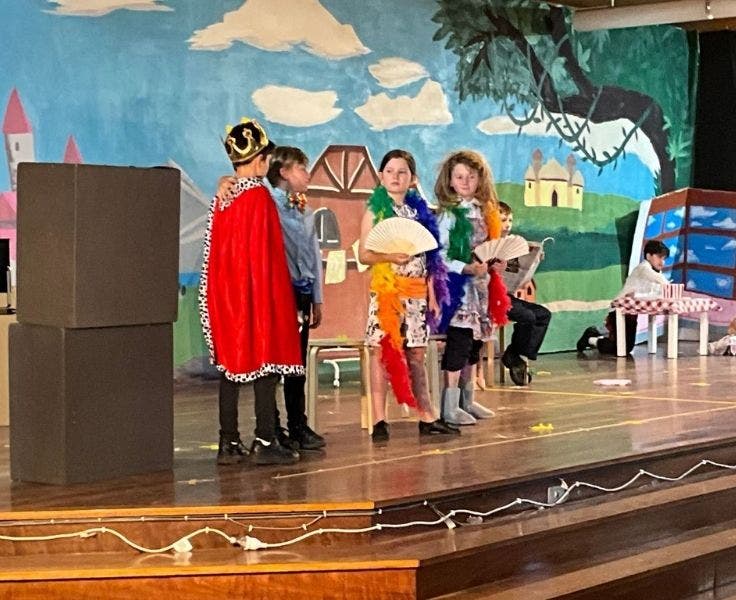 Four students are standing on the stage wearing costumes. There is a fairytale-inspired backdrop behind them.