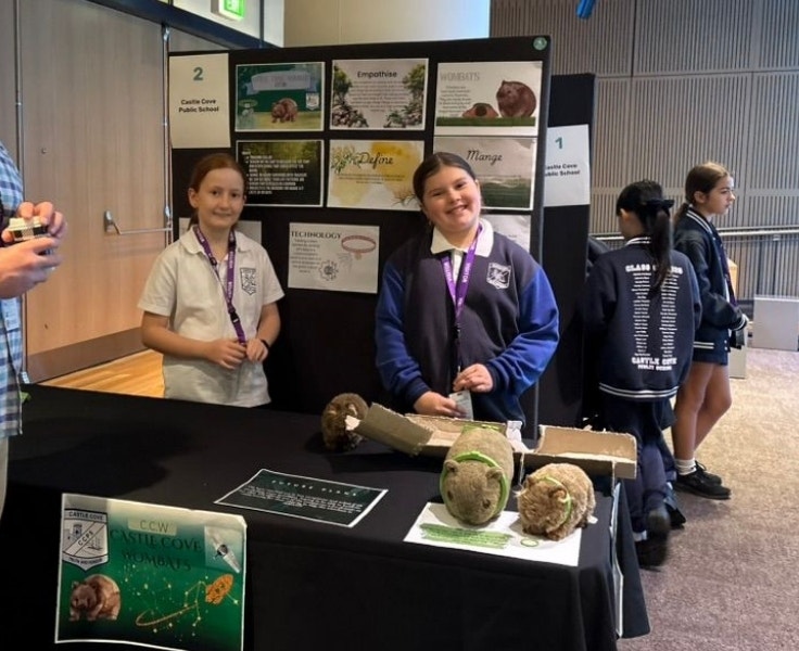 Two female students smiling at the camera as they stand behind a table that has three toy wombats wearing collars on it. There is a board with papers on it behind them.