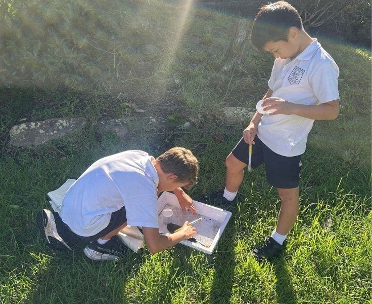 Two boys are conducting an experiment. One boy is crouched down looking at something in a white tote tray while the other boy watches and holds a plastic container.