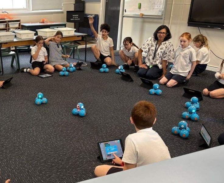 A group of students are sitting around in a half circle with the teacher sitting in the middle of the circle. The students have iPads and blue robots in front of them. One student has his hand raised.