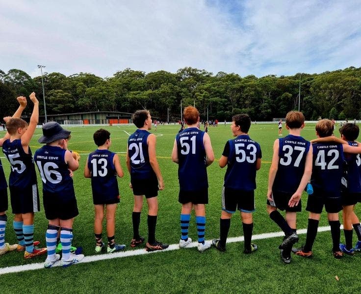 A row of 9 male studnets cheering on an AFL game. They are each wearing a jersey. The jersey numbers from left to right read: 15, 16, 13, 21, 51, 53, 37, 42.