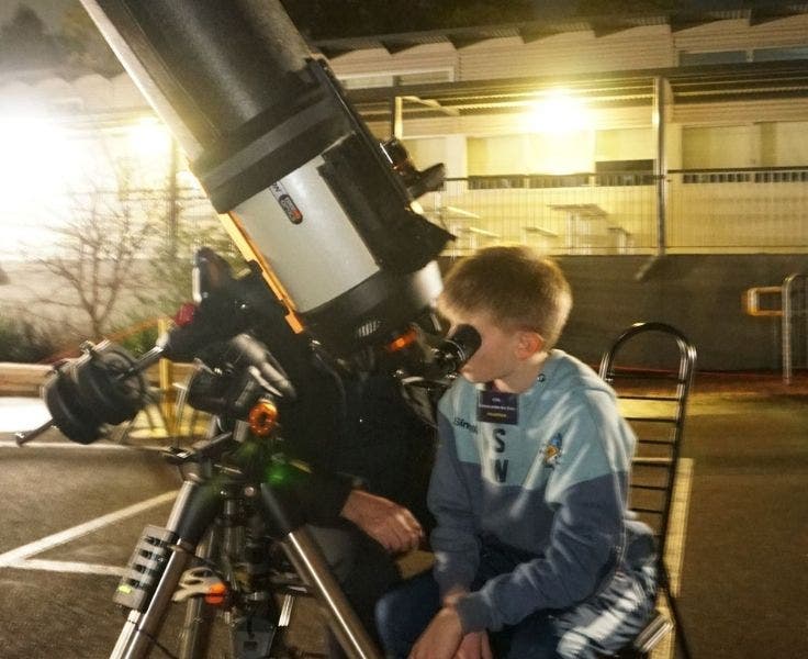 A Year 6 boys is sitting on a chair and looking through a telescope.