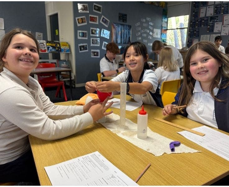 Three female students sitting at a desk. One is smiling at the teacher behind the camera, the other two are smiling at the camera. There is a science experiment happening in the middle of the table.