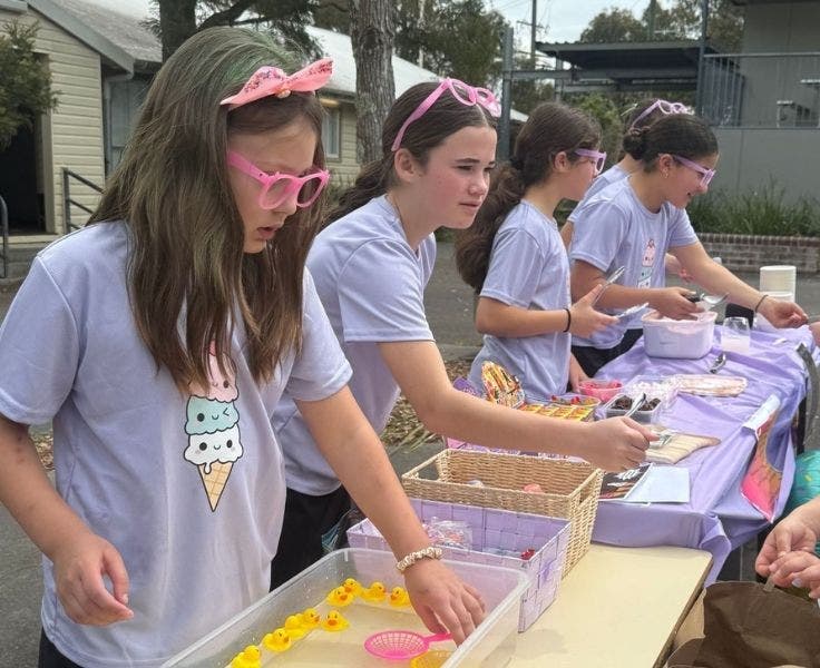 A line of female students wearing lilac shirts are working behind a table.
