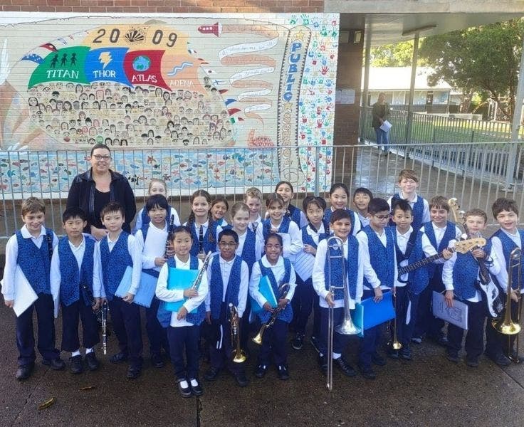 The junior band wearing white shirts and blue blazers are standing with the band conductor. The students are holding their instruments in one and and their music sheet folder in the other.