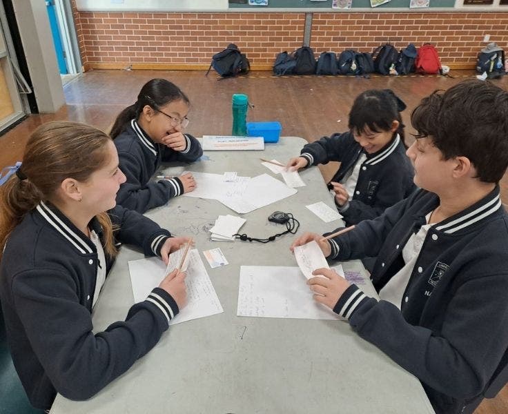Four students sitting at a grey table. They have pieces of paper in front of them. They are laughing.
