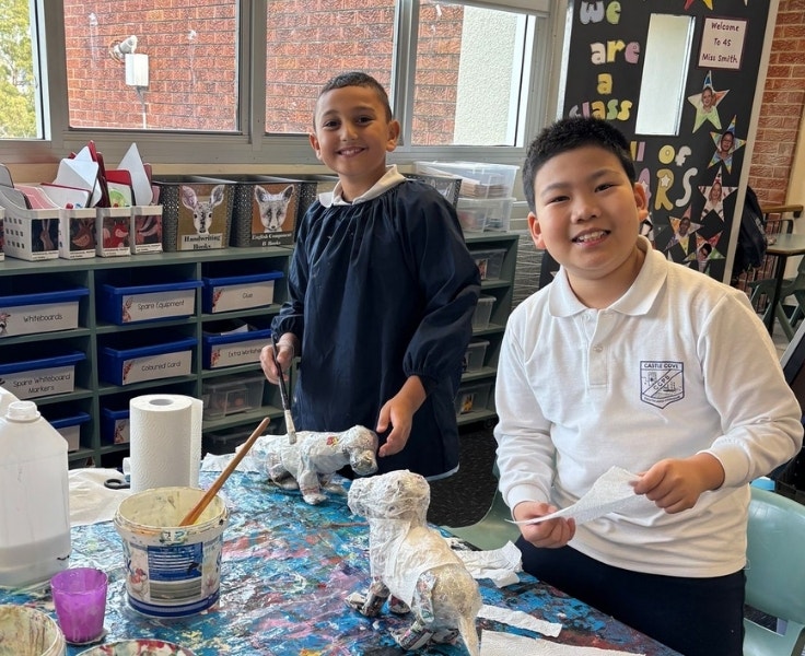 Two male students standing at a table and smiling at the camera. Both boys have a paper mache dog in front of them. One of the boys is holding a piece of paper and one is holding a paintbrush.
