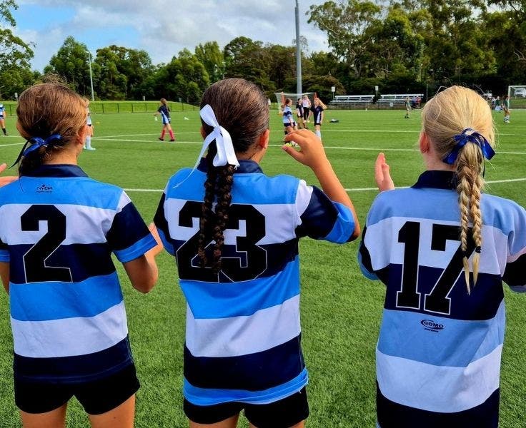 Three girls wearing striped jerseys cheering on a soccer match. One jersey reads