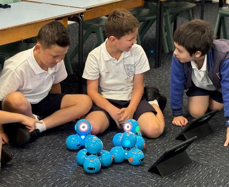 Three male students are sitting on the floor with 4 blue robots in front of them. The boy in the middle and the boy on the right are laughing together.