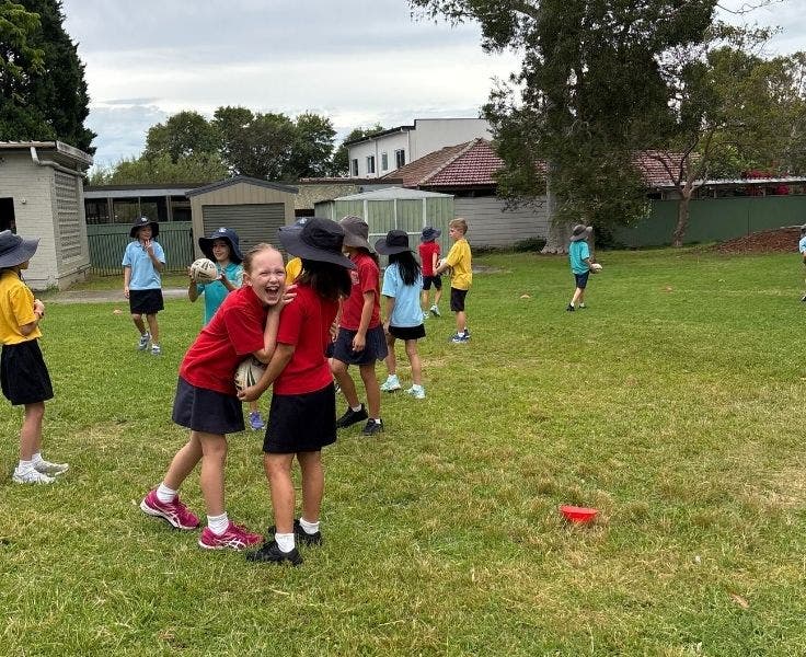 Students wearing a variety of coloured shirts are standing on a field. One female student is holding onto another and laughing in the direction of the camera.