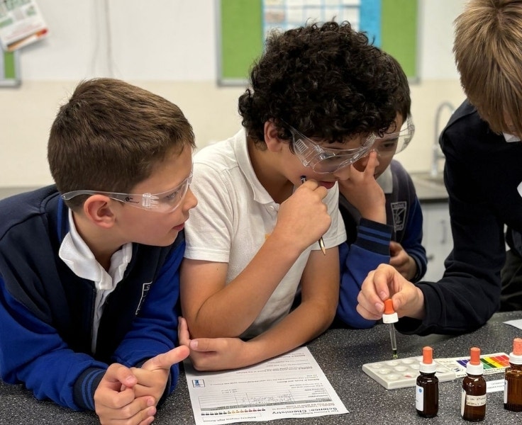 Three male students are wearing safety goggles and watching as an older student uses an eyedropper to put a liquid in a tray.