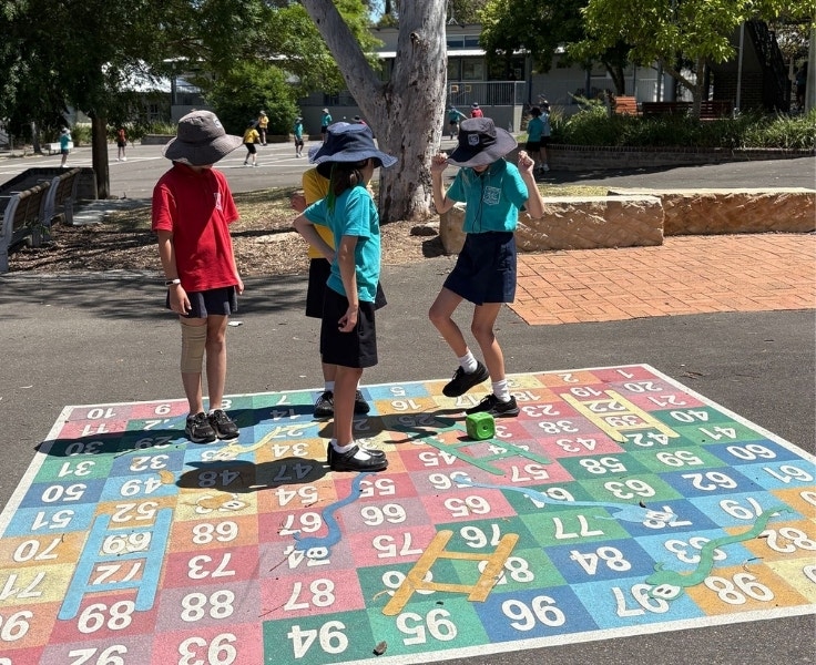 Four students standing on a 1-100 chart. Three of them are watching as the fourth jumps to the next number. There is a green die on the ground.