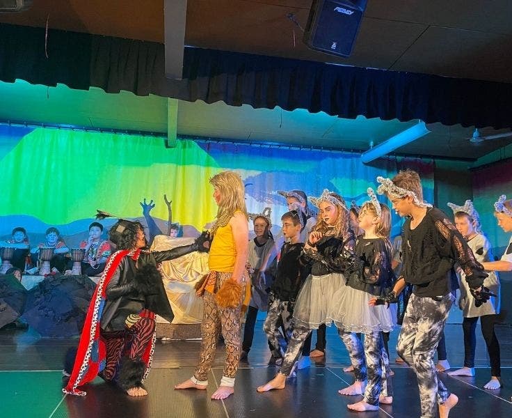 One student is crouched down and holding his hand out to another student who has a group of people behind him. They are all wearing Lion King-inspired costumes.