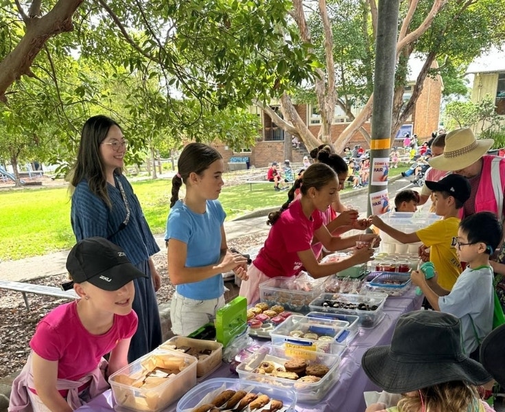 A group of Senior girls are selling baked treats to some younger students as a parent supervises behind them. A teacher in a pink vest is speaking with a student.